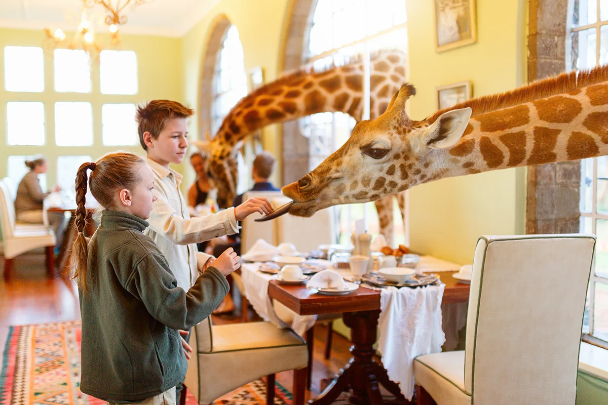 At Giraffe Manor hotel in Kenya, two children feed a young giraffe poking its head through a window