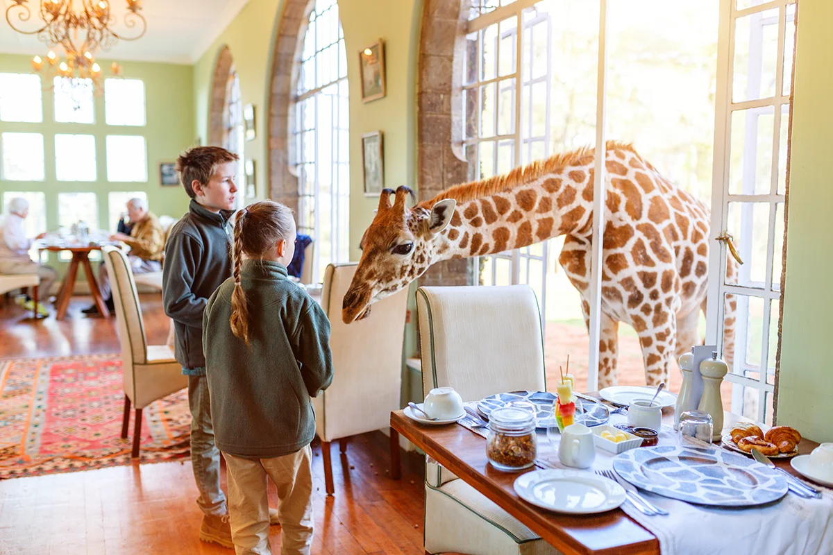 At Giraffe Manor hotel in Kenya, two children feed a young giraffe poking its head through a window