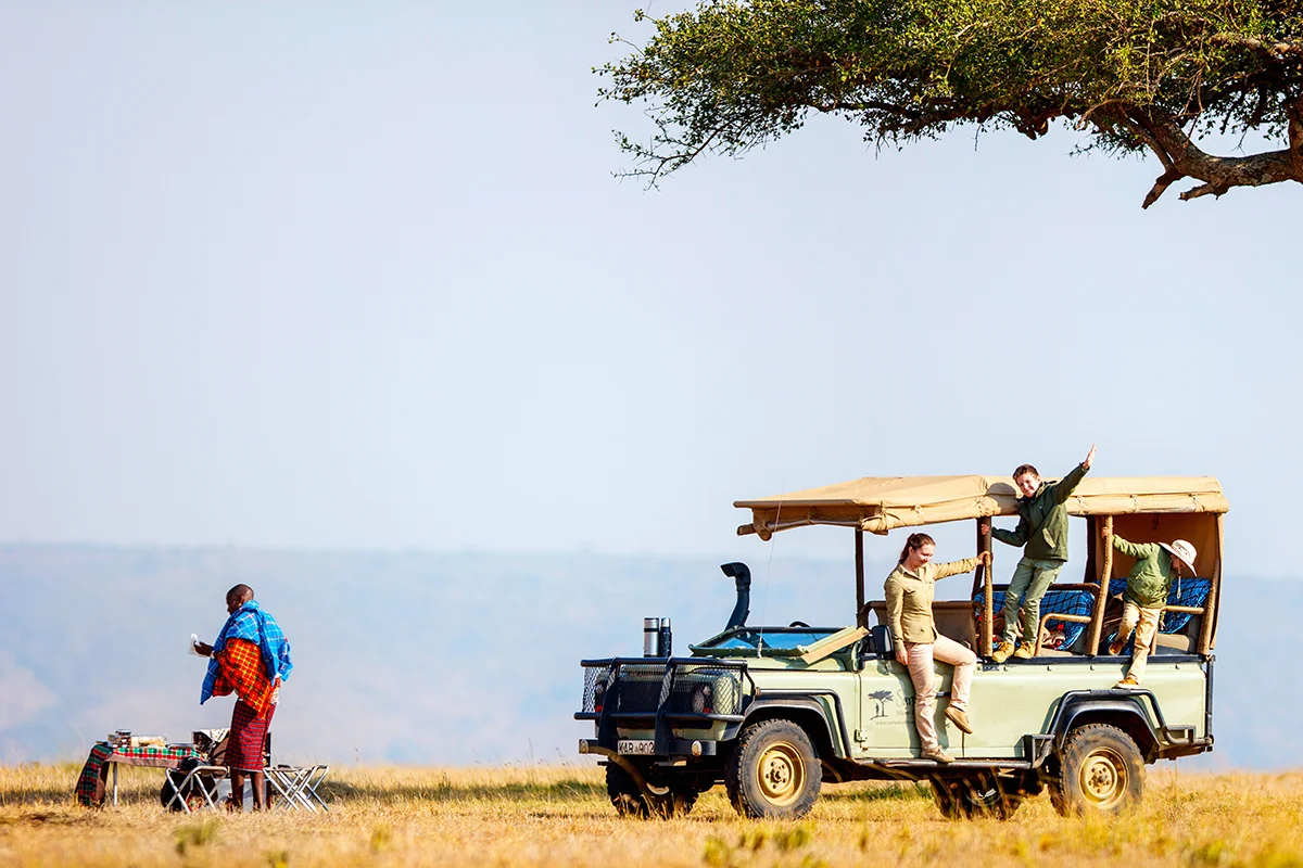Maasai guide setting up a table for a family bush breakfast next to a safari vehicle in Kenya