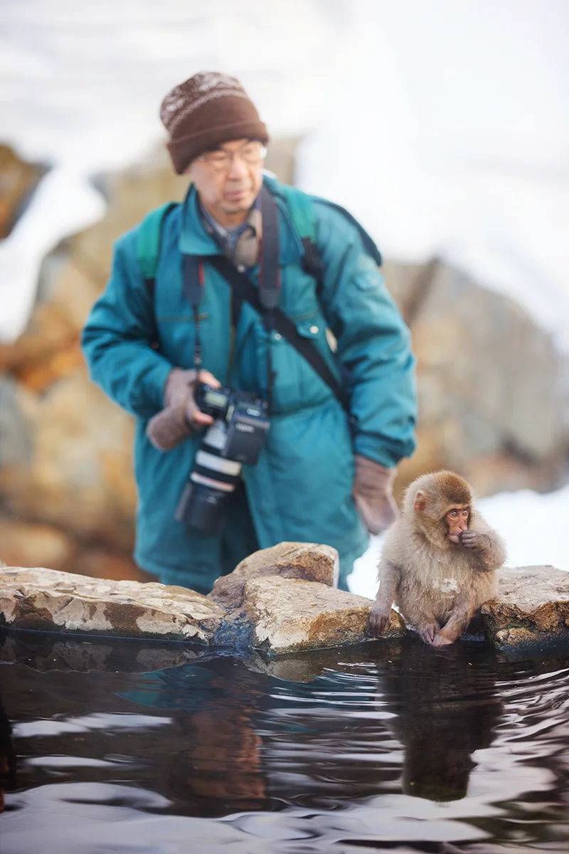 Man photographing monkey by water in Jigokudani Snow Monkey Park, Japan