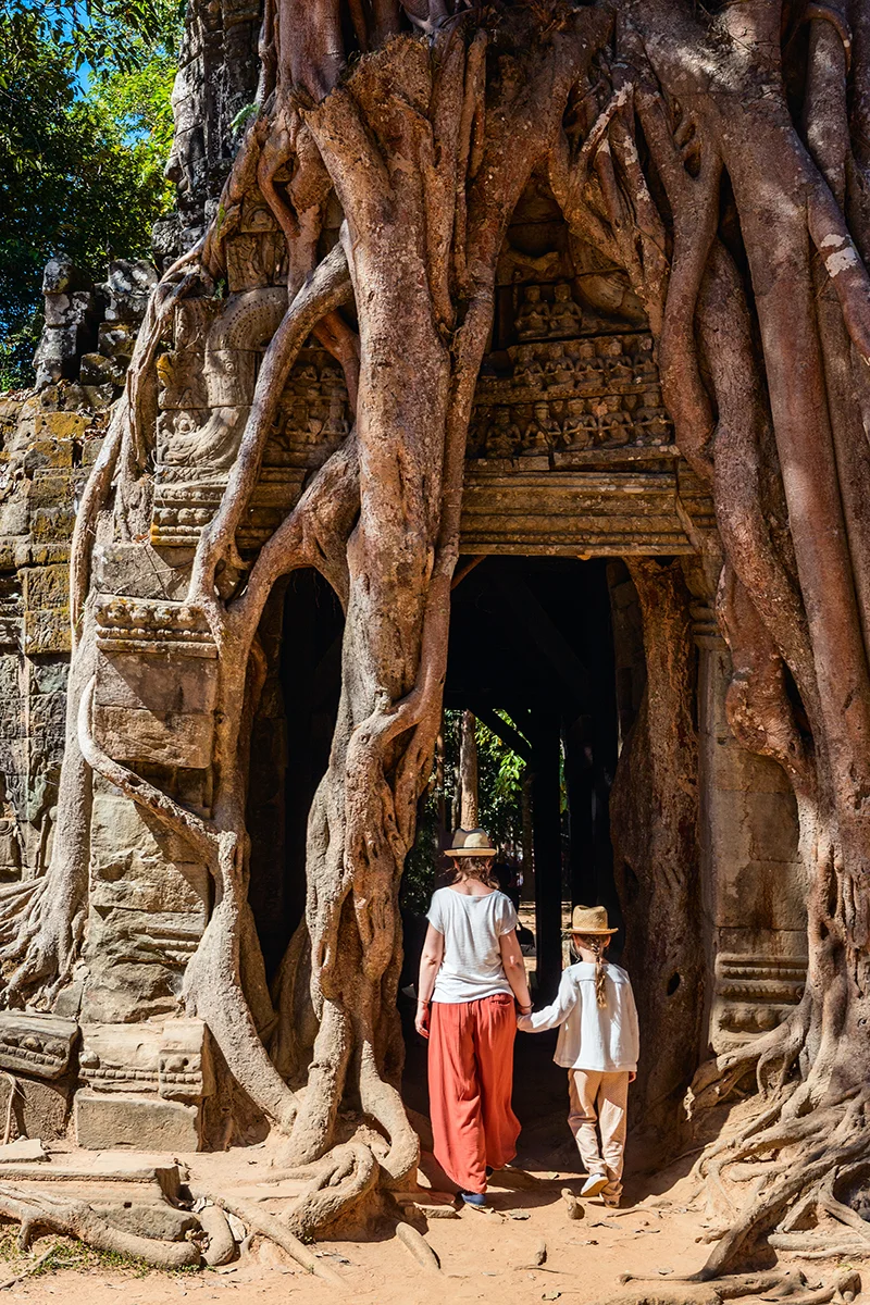 Tourists at iconic entrance of Ta Som temple in Angkor, where massive tree roots dramatically cover the ancient stone gateway